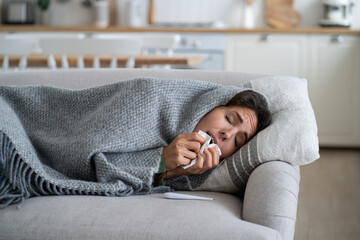 Winter illnesses. Sick female sneezing in tissue while lying under blanket in cold apartment. Young...