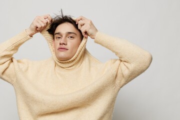 Horizontal studio shot. A beautiful young man with dark short hair combed back in a beige sweater with a high neck stands on a gray background