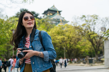 Fototapeta na wymiar stylish taiwanese female photographer wearing sunglasses and holding camera is looking into distance at beautiful landscape in Osaka castle park at springtime in japan