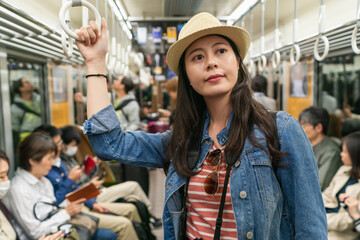 portrait of happy asian girl wearing hat taking Osaka subway with other passengers in japan. she holds onto the handle ring and looks into space with a smile on the train