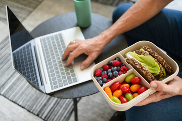 Hand of man holding lunch box with healthy food and using laptop at home