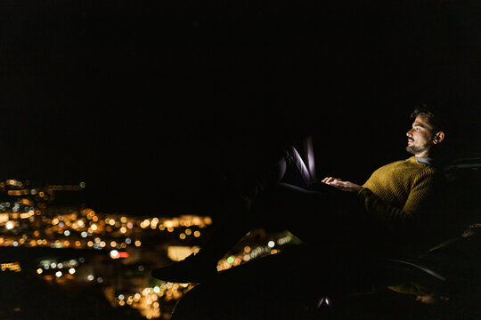 Man sitting on hill above illuminated city at night using laptop