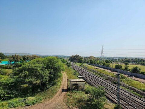 Stock photo of railway track or train pathway surrounded by green trees and plants. Electric poles and small old cabin near the railway track. Picture captured under bright sunlight at Gulbarga, India