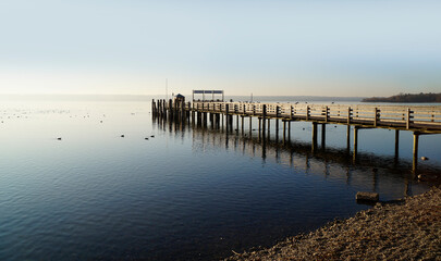 Obraz premium a long wooden pier in Herrsching on calm lake Ammersee in Bavaria on a clear serene January evening (Herrsching, Bavaria, Germany) 