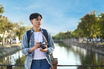 Young asian male blogger with digital camera standing on bridge with scenery view of river and beautiful blue sky