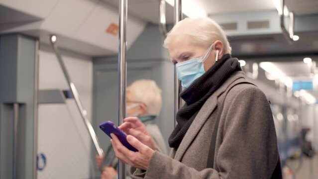 women in a protective mask using a smartphone standing in a subway car.