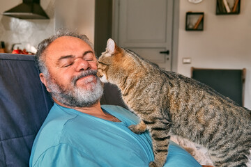 Tabby cat licking face of bearded man in living room. Human-animal relationships. Pets care. Funny home pet. Cat day. Selective focus. Adopted pet. - obrazy, fototapety, plakaty