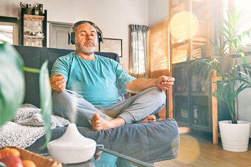 Mature middle-aged overweight man in wireless headphones relaxing at home with guided meditation, listening to relaxing music on smartphone and meditating in lotus pose.