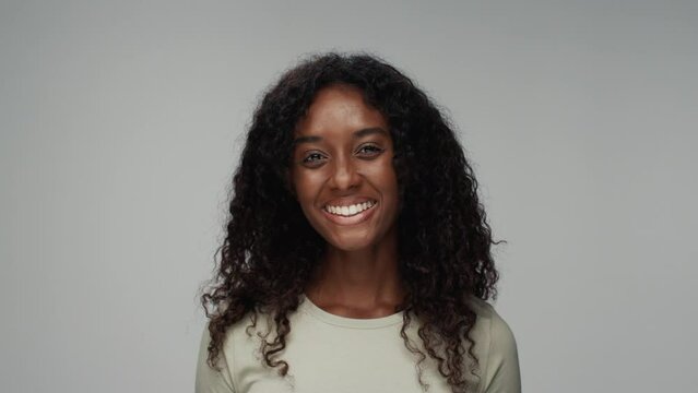 Portrait of Young Black Woman Looking at Camera and Smiling in Colour Studio Shot. Happy Cheerful Girl Isolated Alone on Grey Background Closeup. African American Person with Healthy Face Turning Head
