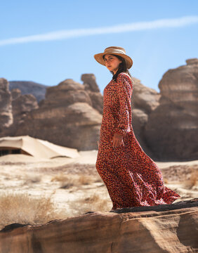 A Girl wearing a long red dress and a straw hat stands up on top of volcanic rocks in Al Ula, Saudi Arabia