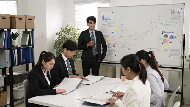 asian male mentor coach wearing suit is hosting a trainee seminar in the meeting room. he asks all interns to open the folder and read some document
