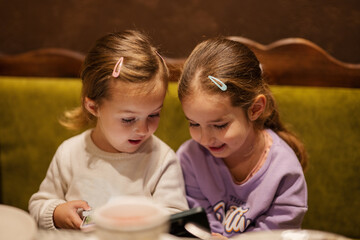 Sisters kids hold mobile phone together at cozy cafe and having fun.