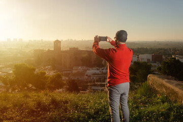 Man in sportswear on top of a hill taking a picture of the landscape with his mobile phone with a medieval village in the background