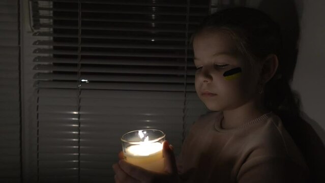 A girl with a painted flag of Ukraine on her cheek in a dark room holds a candle in her hands