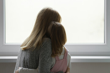 Young adult mother and daughter together looking out from window at home. Waiting, longing or...