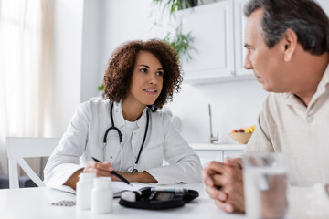 cheerful african american doctor pointing with pen at glucometer device near middle aged man with diabetes