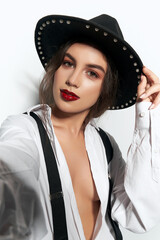 Close-up shot of a woman in a fedora hat adorned with decorative pearls. The lady in a white unbuttoned blouse, black suspenders and the felt hat with pearl beads is posing on a light background.