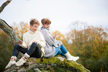 Fototapeta premium Happy mother and son teen sit with their backs together on a tree branch and use cell phones and headsets