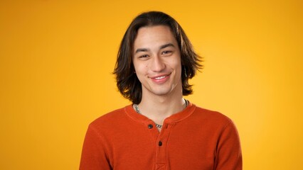 Smiling handsome Hispanic young man 20s wearing orange casual shirt isolated on yellow color background in studio. Portrait of gender fluid male sincere emotions lifestyle concept. Looking camera
