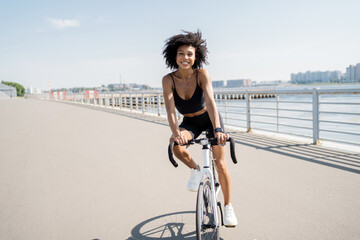Resting and relaxing, a millennial cyclist rides on environmentally friendly transport in the city. A curly-haired happy woman rides a bicycle. - obrazy, fototapety, plakaty