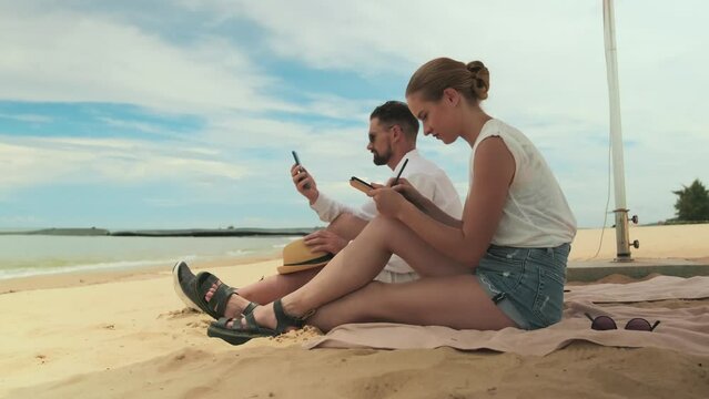 Young creative woman taking a look at seaside while sitting on sandy beach and drawing it on tablet screen against man with smartphone taking selfie or photo of sea panorama during summer vacation