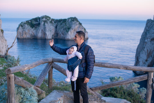 Father with small toddler girl in carrier baby sling. Travel with child. Man make picture selfie with beautiful sea view, talking by video on phone. outdoor