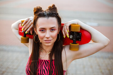 Young female skater with penny board in hipster outfit looking at camera in city in summer 