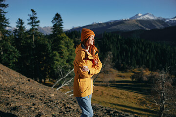 Woman full-length hiker smiling with teeth standing frozen on the mountain looking at nature happiness mountains and trees in yellow raincoat travel and hiking in the mountains at sunset freedom