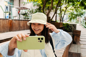 Smiling asian girl taking selfie with mobile phone while sitting outdoors