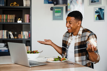 Positive african man having video call via laptop and gesturing while having lunch