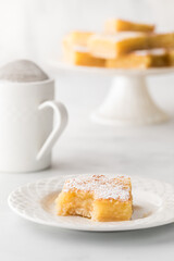 A lemon square with a bite out on a plate and desert stand with more in behind.
