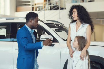 Black salesman asks female customer daughter opinion about cars in automobile salon. Black-haired mother and girl laugh and try to choose car together