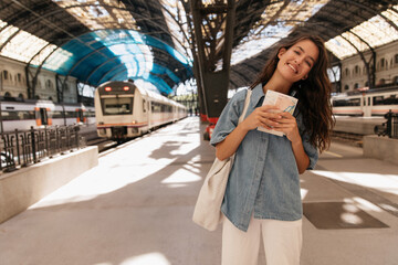 Positive young caucasian girl looking at camera with wide smile holds map in hands. Woman with brunette hair wears jeans jacket and casual jeans. Concept of happy weekend, vacation.