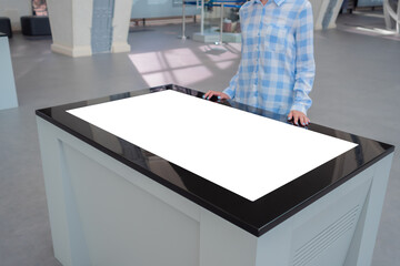 Woman in blue plaid shirt looking at blank interactive touchscreen white display of electronic table kiosk at exhibition or museum. White screen, mock up, copyspace, template, technology concept.