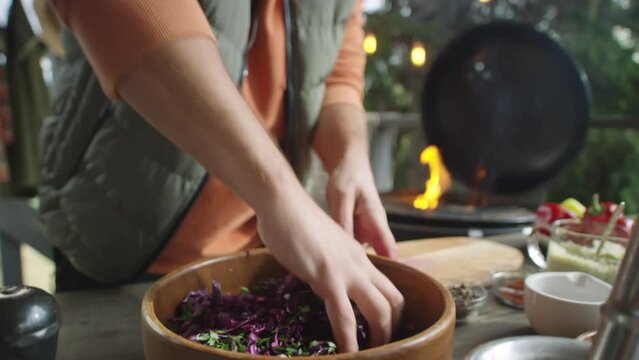 Food blogger mixing salad with hands in bowl and talking on camera while filming cooking vlog on outdoor barbecue terrace