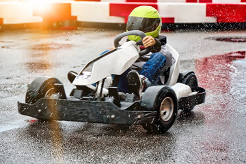 Little racer on the map races on wet asphalt in a protective helmet, splashing from puddles. Young...