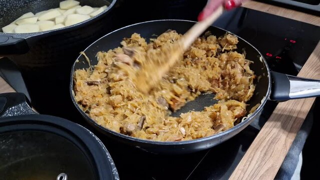 Woman is preparing traditional Polish dish bigos (hunter`s stew) with sausage, mushrooms, meat and cabbage on a frying pan in kitchen