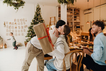 Happy family exchanging gifts during dinner at table while son playing near Christmas tree