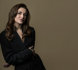 Woman with long dark wavy hair and black suit studio portrait on beige background. Sexy looking woman with day make-up looking at camera with seductive look.Toned image with dark blue color