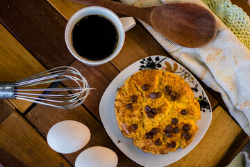 Delicious cornbread with chocolate chips, placed in a white porcelain dish and composed with eggs, wooden spoon, whisk, white cloth and a cup of coffee