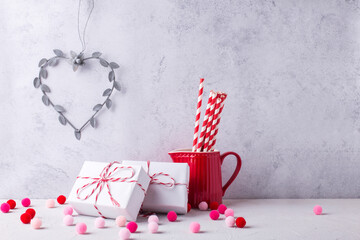 Postcard with  wrapped presents, heart, paper straws in red pitcher, pink and red pom poms against  grey  textured  wall.  St. Valentines day postcard. Place for text.