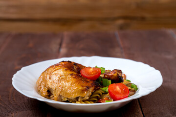Pasta with vegetables and baked chicken leg with tomatoes sprinkled with green onions in a plate on the table.