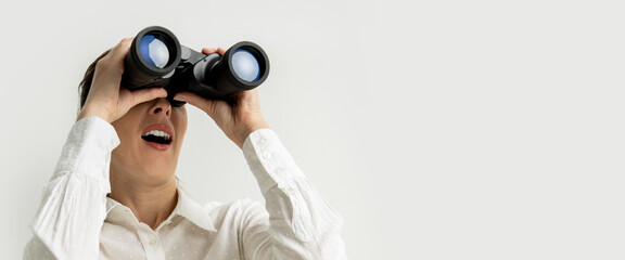 Surprised young woman in a white shirt looks through binoculars on a white background. Banner