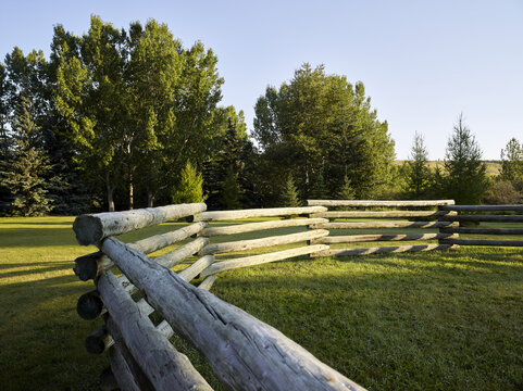 Close-up of an empty paddock with a split rail fence on a grassy field surrounded by trees with shadows from the sunlight, Historic Cochrane Ranche Park; Cochrane, Alberta, Canada