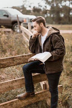 A young man stands holding his open Bible beside a rail fence and truck park on a road in the countryside; Alberta, Canada