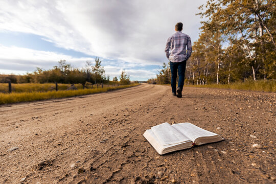 A young man walks down a country road away from Bible laying open;  Alberta, Canada