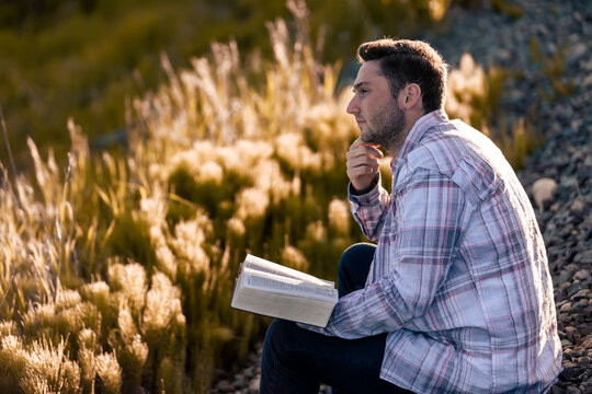 A young man reads from his Bible as he sits in the countryside; Alberta, Canada