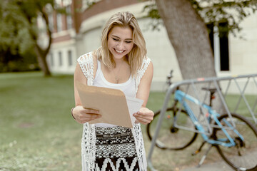 A female university student stands outside on campus looking at a document out of an envelope and receives good news; Edmonton, Alberta, Canada