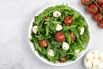 Green salad with arugula, cherry tomatoes, mozzarella cheese and spices in a white plate on gray background. 