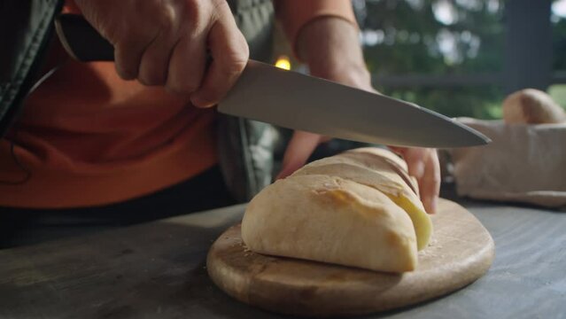 Cropped shot of man cutting ciabatta bread with knife on wooden board while cooking on outdoor terrace